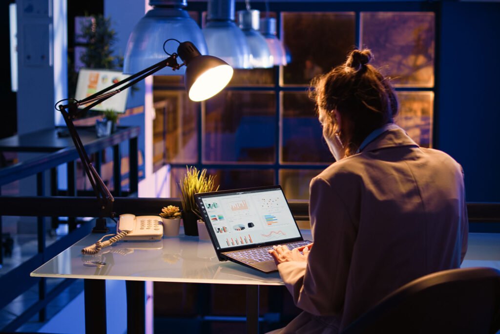 Woman working late at a modern office desk with data analytics on laptop screen